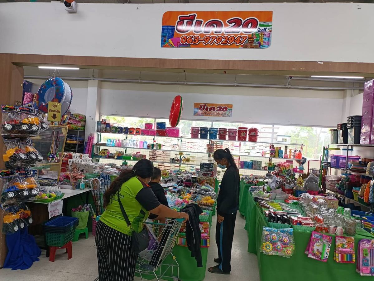 หน้าร้านบีเค20 ป้ายชื่อร้านสีส้มเหนือทางเข้า ด้านในมีของใช้และของเล่นวางเต็มโต๊ะ