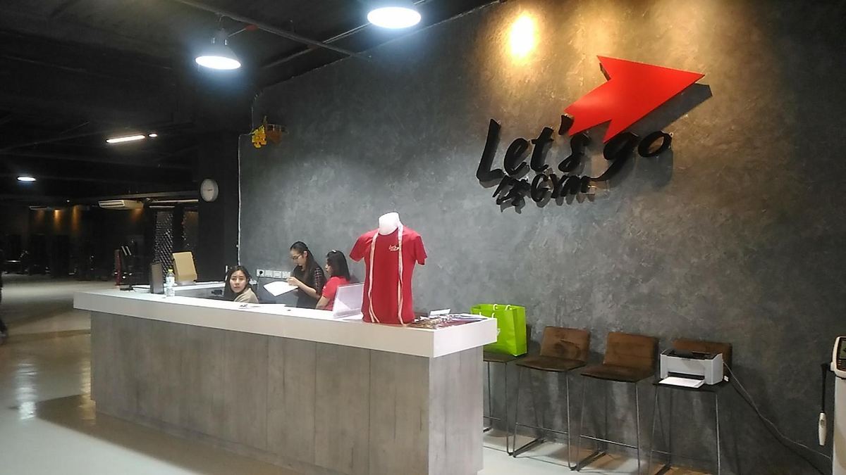 เคาน์เตอร์ต้อนรับของ Let's Go Gym ผนังปูนสีเทาติดโลโก้ลูกศรสีแดง