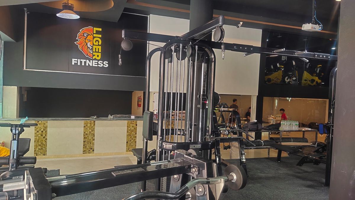 โลโก้หัวสิงโต LIGER FITNESS บนผนังสีดำ ด้านหน้ามีเครื่องเล่นเวทแบบสายเคเบิล