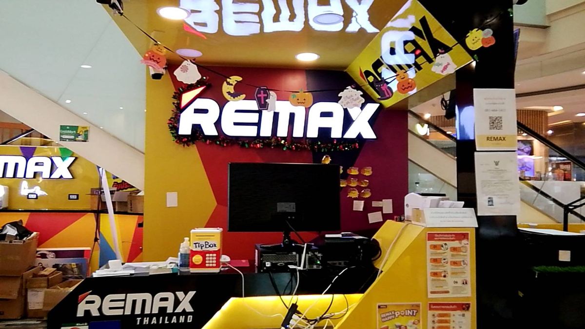 เคาน์เตอร์ร้าน REMAX ในห้าง ป้ายไฟ REMAX สีขาวบนผนังสีเหลืองแดง พร้อมจอคอมพิวเตอร์ที่เคาน์เตอร์