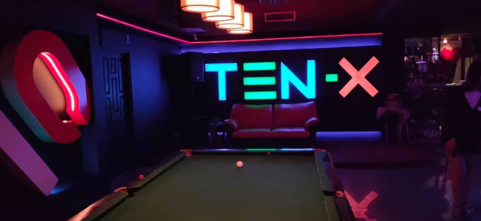 ภายใน TEN-X Karaoke มีโต๊ะพูลหน้าป้ายไฟนีออน TEN-X สีฟ้าและชมพู พร้อมโซฟาสีแดง