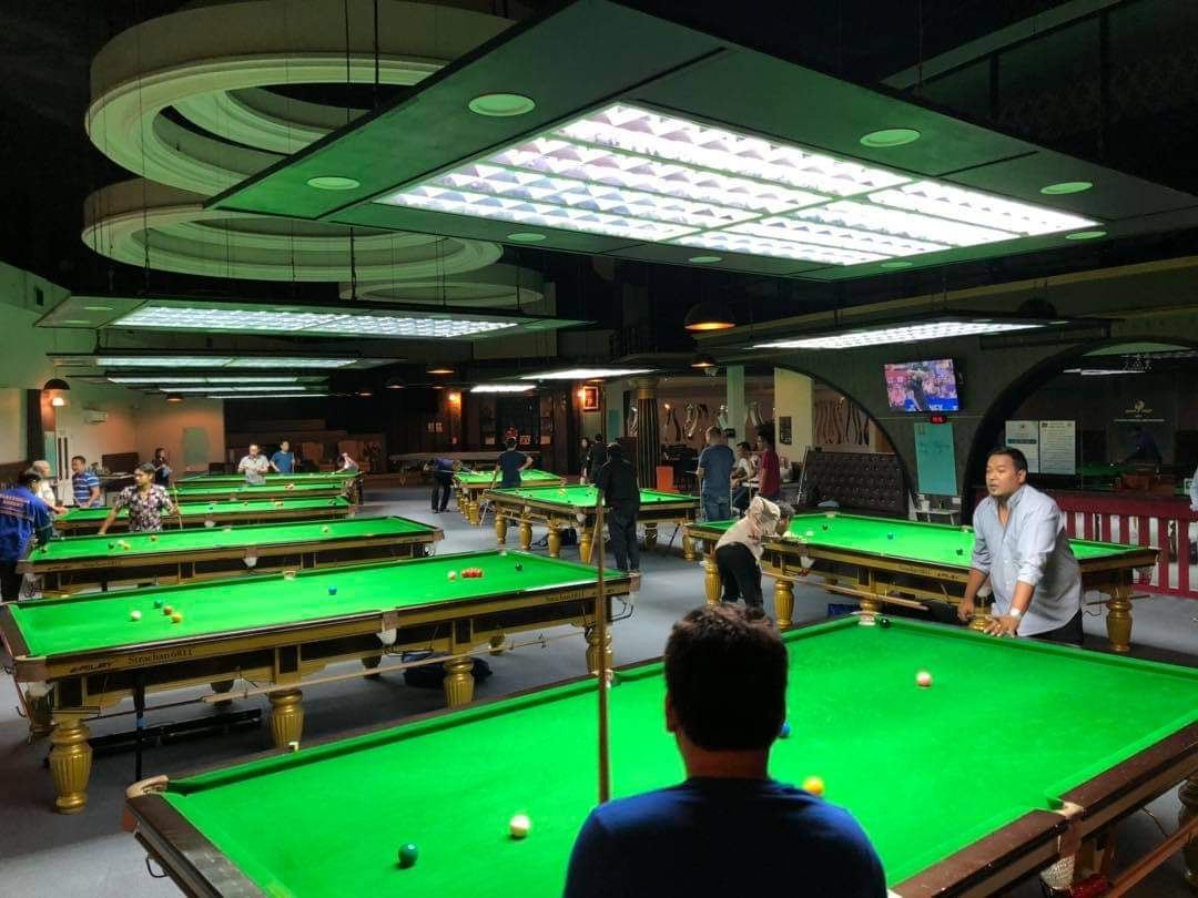 ห้องโถงของ World Class Snooker Club มีโต๊ะสนุกเกอร์สีเขียวหลายแถวใต้โคมไฟ และผู้เล่นกำลังเล่น